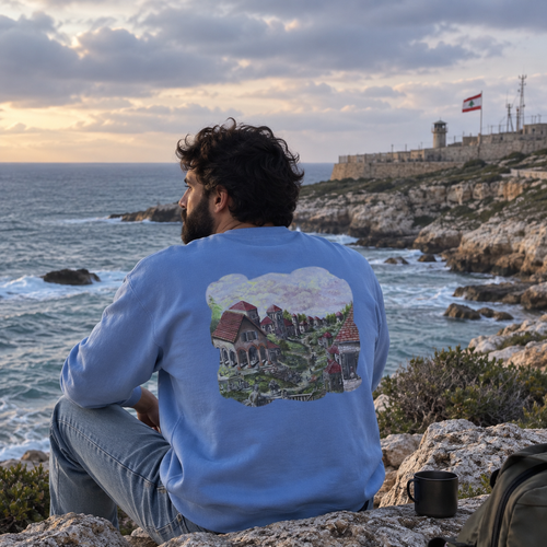 A man sits on a rocky cliff overlooking the ocean, seen from behind. He is wearing a light blue crewneck sweatshirt with a large graphic print on the back depicting a coastal village scene.