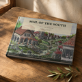 A hardcover book titled "SOIL OF THE SOUTH" lies on a rustic wooden table, angled slightly to the side. The cover features a detailed vintage-style painting of a Southern village scene with red-roofed houses, green pathways, and small figures.