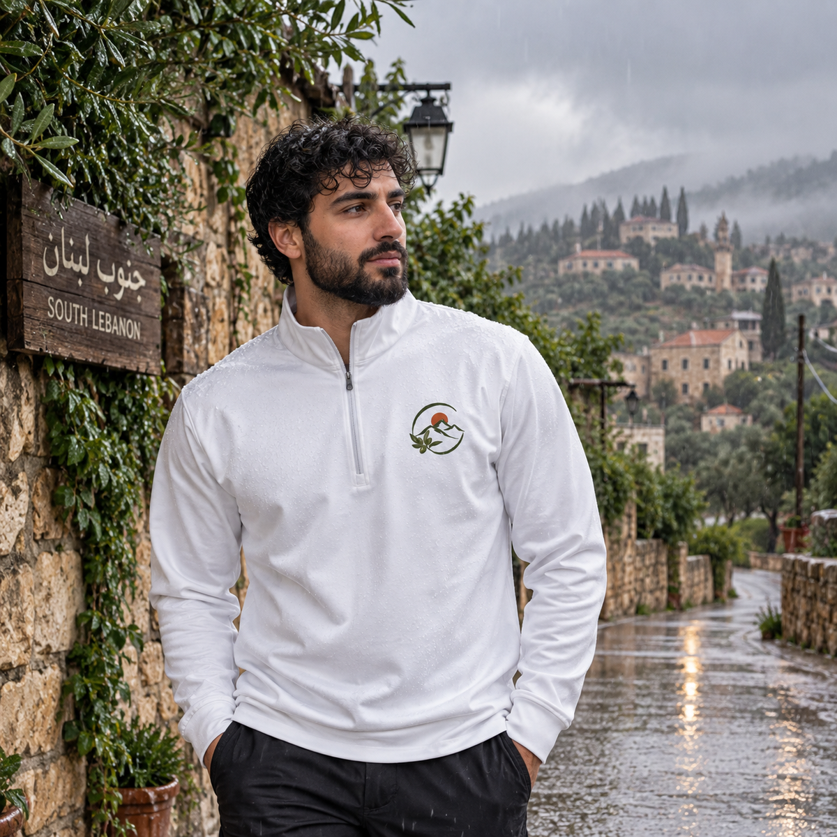 A man wearing a white long-sleeve quarter-zip pullover with a small mountain logo on the left chest, standing on a wet stone street in a village with mountains in the background.
